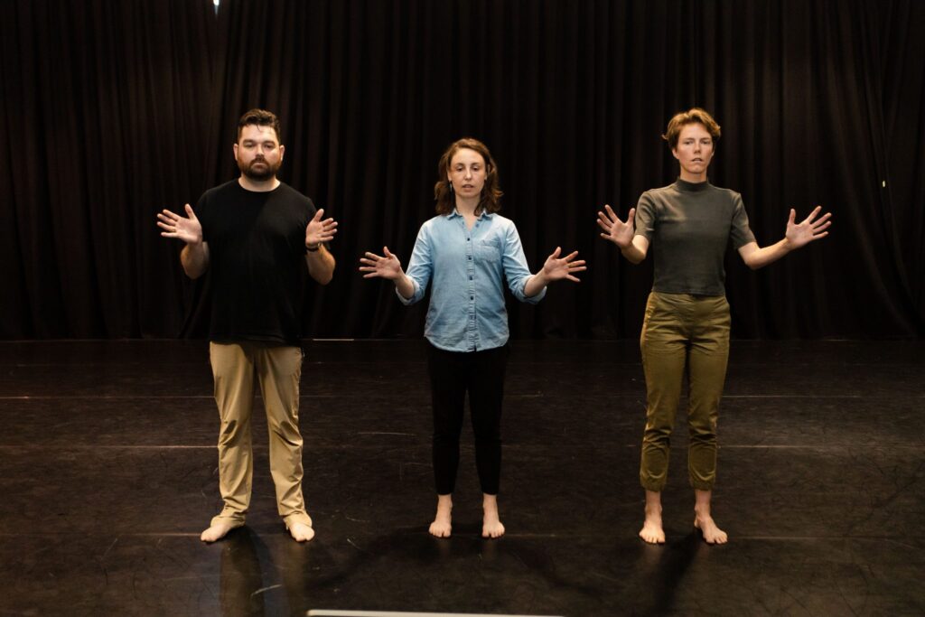 Benedict Carey, Patricia Wood and Cloe Mees standing side by side with open palms on their side in the Drill Hall