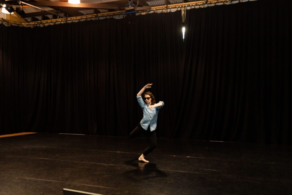 Patricia Wood in the Drill Hall dancing. Patricia is wearing sunglasses, a blue shirt and black pants.