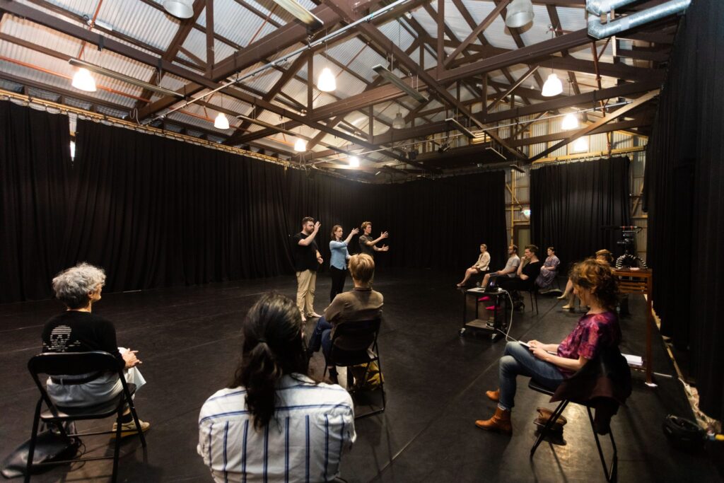 Benedict Carey, Patricia Wood and Cloe Mees in the background of the image, with a small audience in the foreground of the image. The audience are seated on black chairs and are seated with plenty of space between them for COVID-safety.