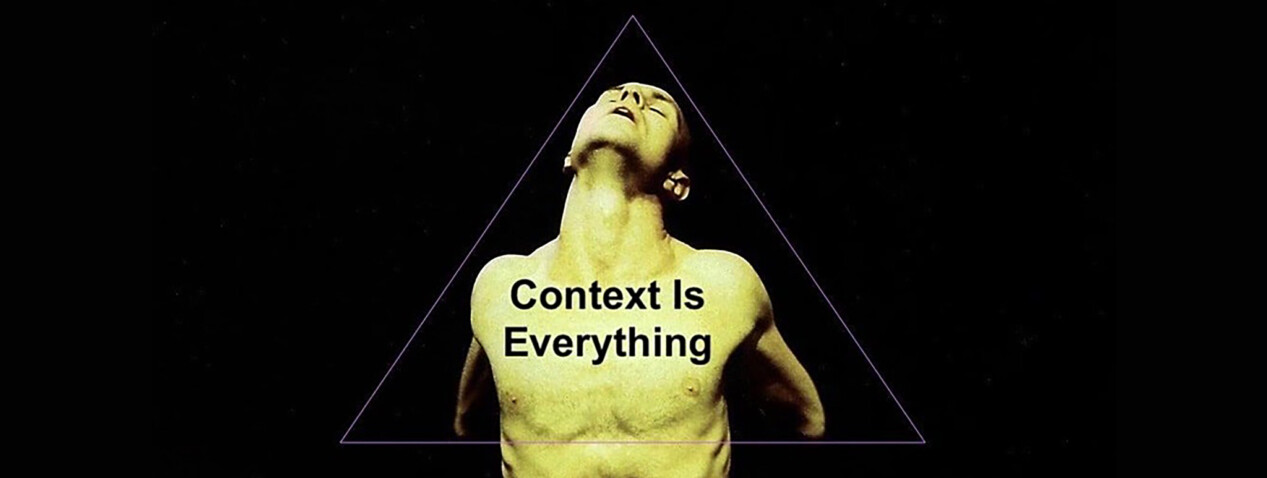 Artist Dean Walsh, topless and looking up into the sky with his arms stretched behind him. His body is against a black background. The words "Context is Everything" is printed across his chest, and there is a pink triangle outline that surrounds his body.