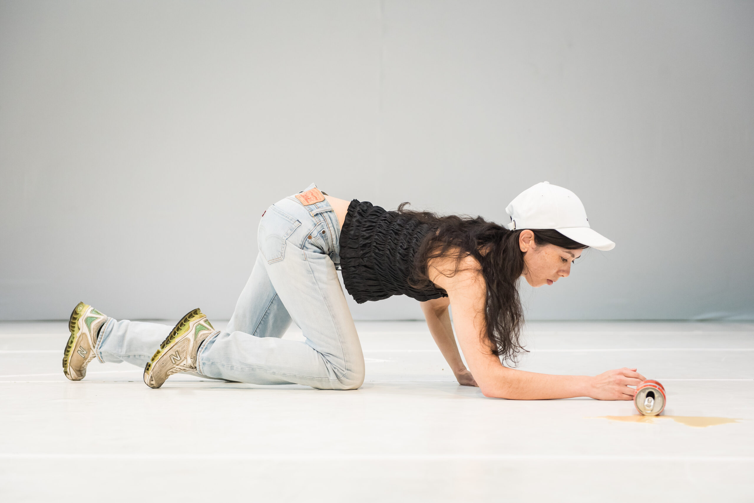 This is a photograph of a woman who is wearing a black sleeveless top, light blue jeans, brown sneakers and white hat. She is crawling on the floor and there is a spilt can of Coca Cola in front of her.