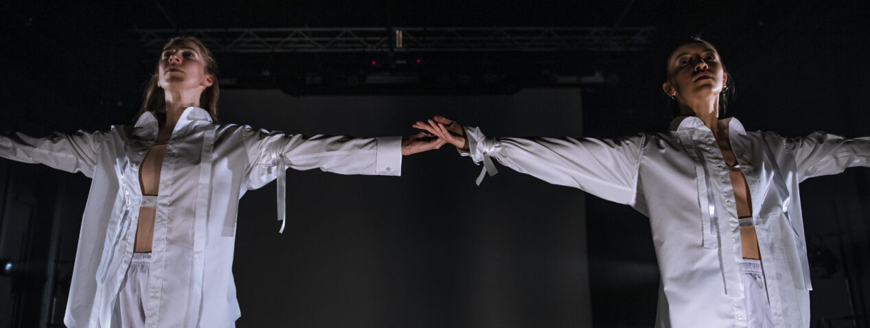 Two dancers in a white long-sleeved shirt and shorts stand next to each other with arms outstretched. Both look into the distance.