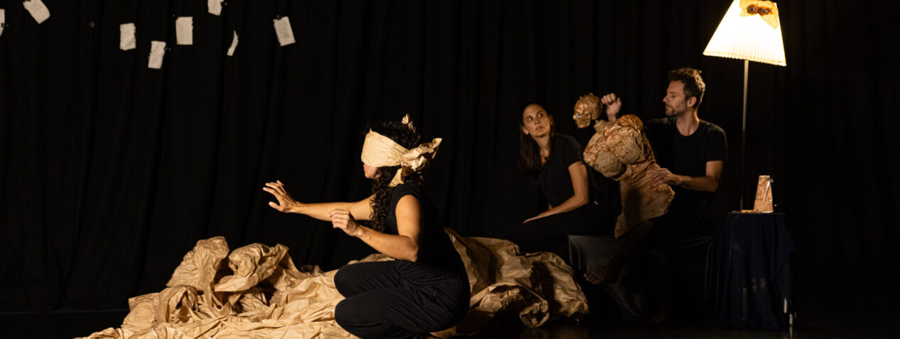 ID: A blindfolded woman in a dimly lit room crouches on the ground and reaches out into empty space. There are two figures seated in the background. One of them grapples with the torso of a life-sized puppet.