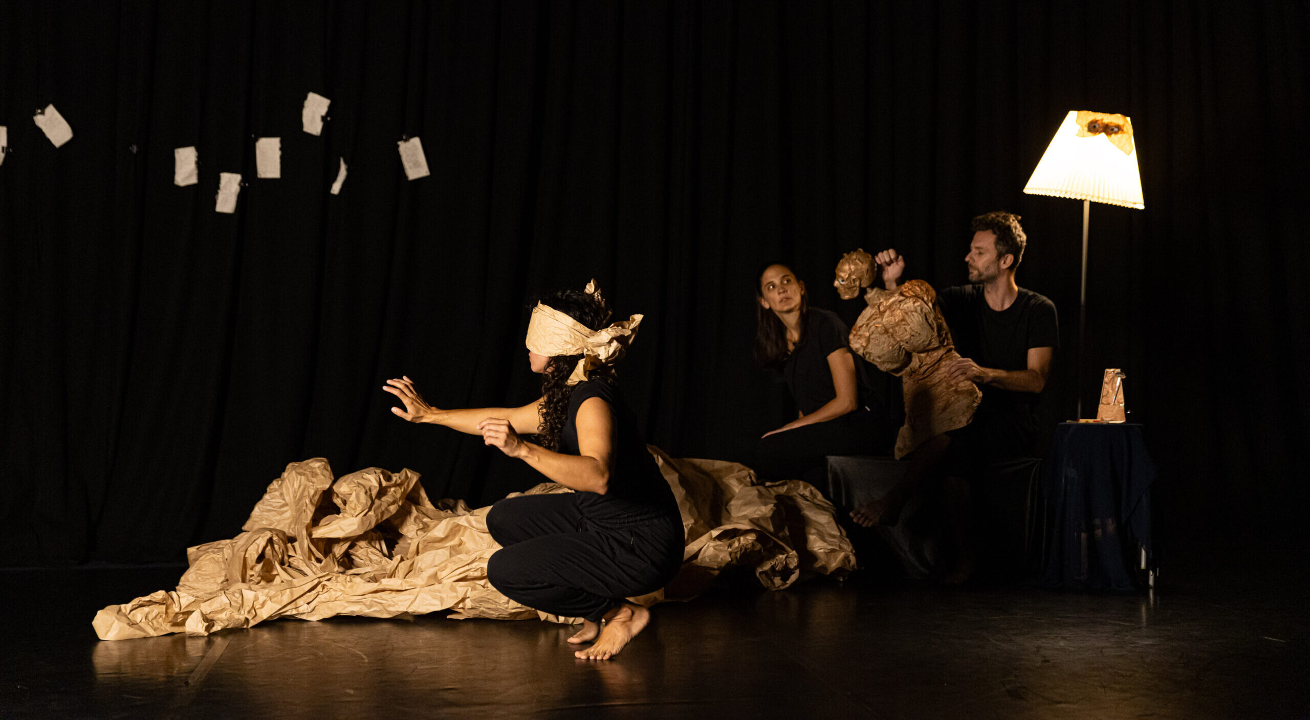 ID: A blindfolded woman in a dimly lit room crouches on the ground and reaches out into empty space. There are two figures seated in the background. One of them grapples with the torso of a life-sized puppet.