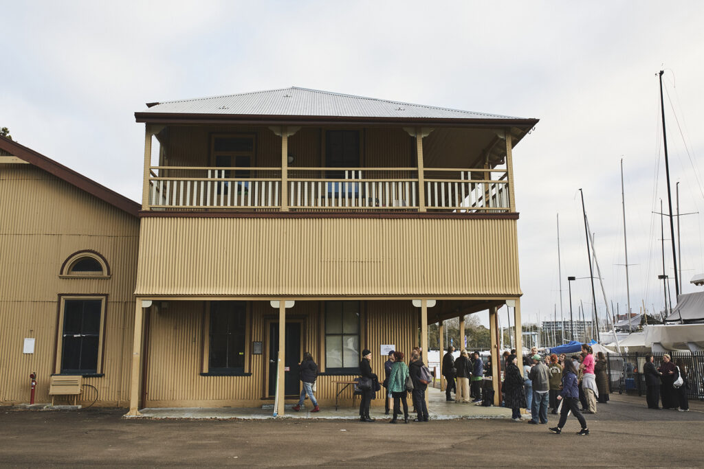 A day image of The Drill Hall in Darling Point, Sydney. It is a two-storey, mustard heritage building. People are gathered outside The Drill Hall