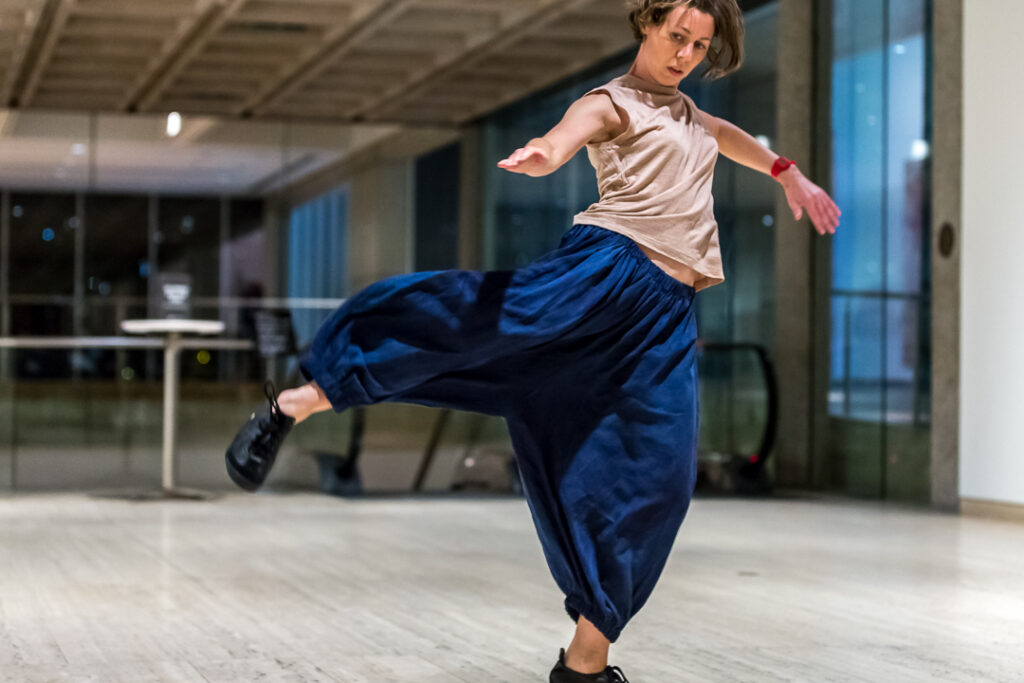  Image description: Photograph of dancer in gallery. Her leg is outstretched and she is looking at it. 