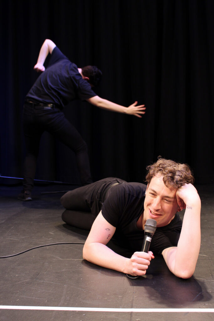 Photograph of two non binary performers wearing black T-shirts and jeans. The performer in the background, on the left side of the image, is turned away mid movement.The performer in the foreground is lying on their stomach speaking into a microphone.