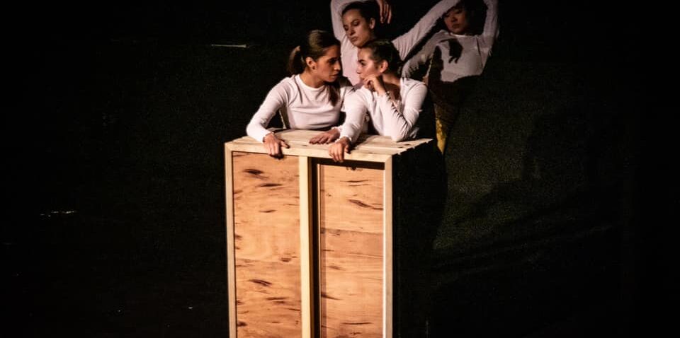 ID: Photograph of four girls standing on a black floor, wearing white shirts. They are having a conversation behind a square wooden structure with wheels.