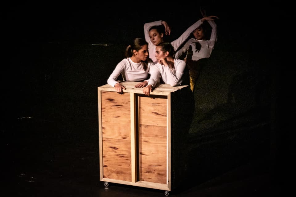 ID: Photograph of four girls standing on a black floor, wearing white shirts. They are having a conversation behind a square wooden structure with wheels.