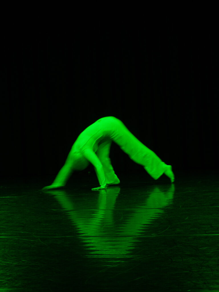 ID: A photograph of a female dancer walking on her hands in feet in the centre of a dark performance space. The dancer is green from the lighting and is softly in focus