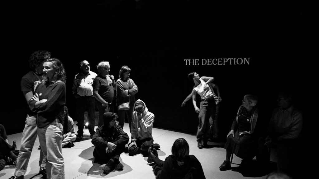 Black and White image of two dancers performing near a wall with the words 'The Deception' projected onto it.