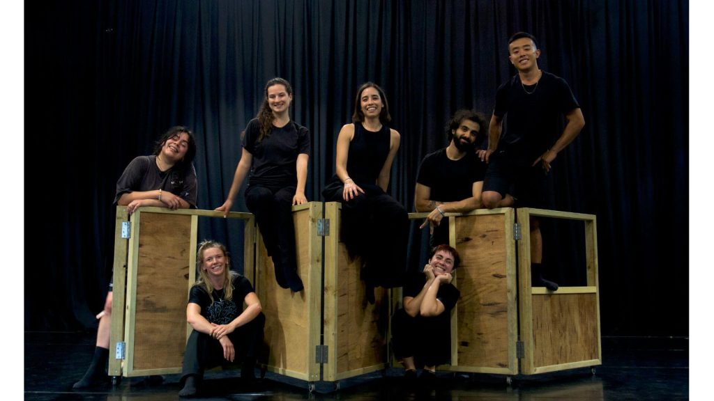 A photograph of seven performers sitting on top of a wooden structure with wheels. They are all wearing black outfits, smiling and looking directly at the camera.