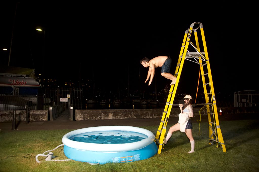 A photograph of a scene taken at night on the foreshore of Sydney harbour. A person is mid-flight leaping off a yellow ladder into a small portable baby pool while another person stands holding the ladder still.
