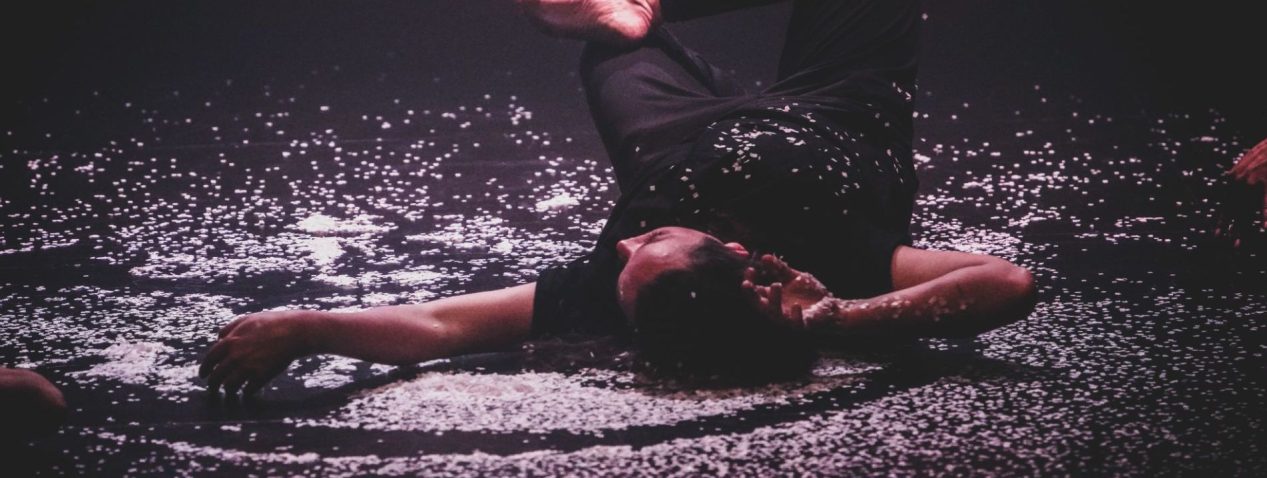 a person lying on their back on the floor of a performance space surrounded by ash.