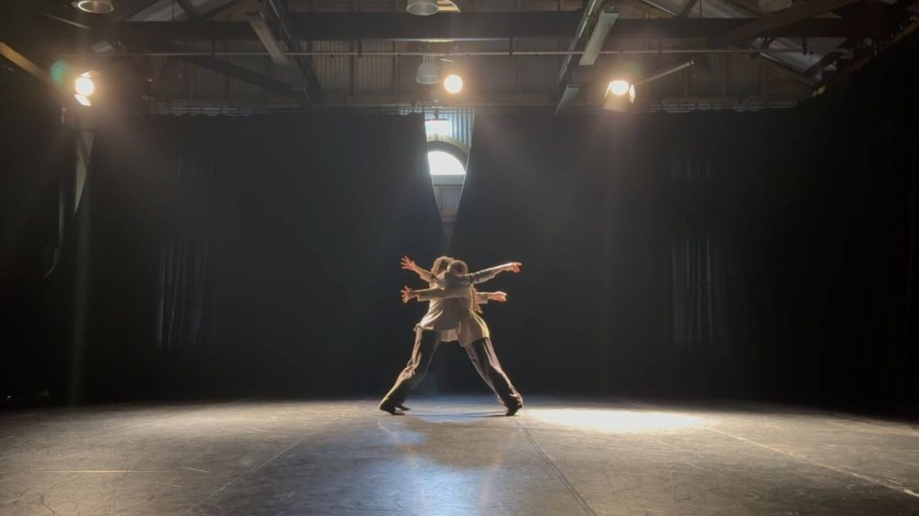 Two performers wearing blazers and black pants in a large room with black curtains and flooring, backlit. They balance against each other chest to chest, with arms outstretched beyond the other.