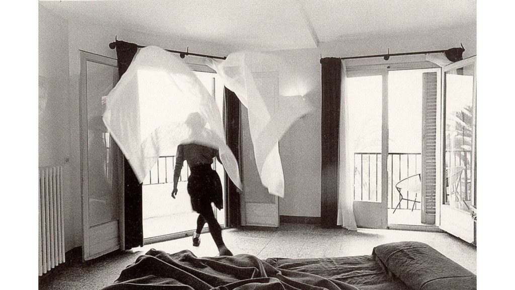 Black and white still of a female figure behind a billowing curtain in a hotel room.