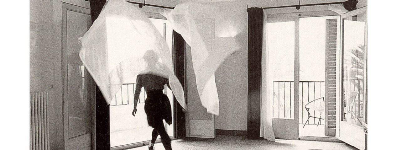 Black and white still of a female figure behind a billowing curtain in a hotel room.