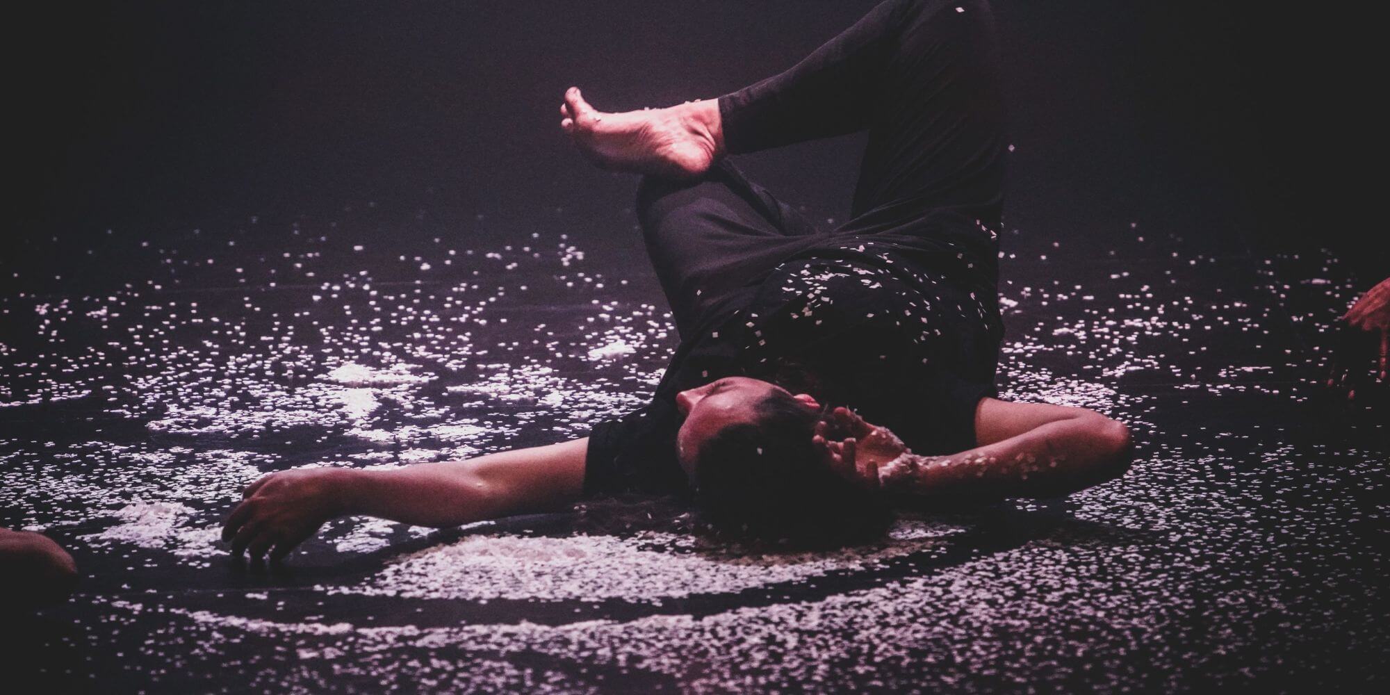 A person lying on their back on the floor of a performance space surrounded by ash.