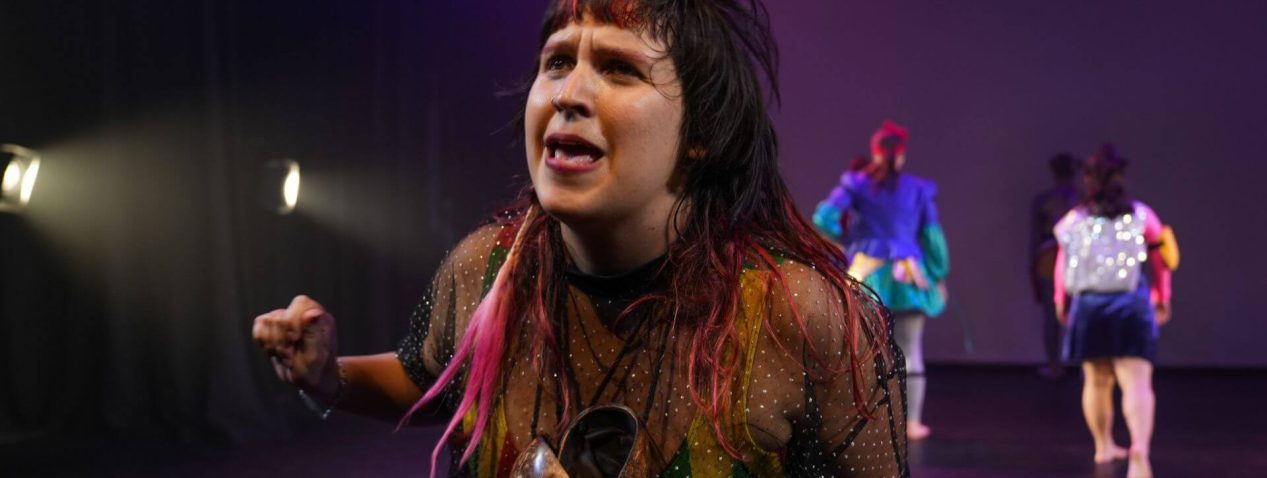 an image of a person on a mauve-lit stage, speaking expressively to the audience, wearing a coconut shell as a necklace and brightly coloured swimmers underneath a sheer top.