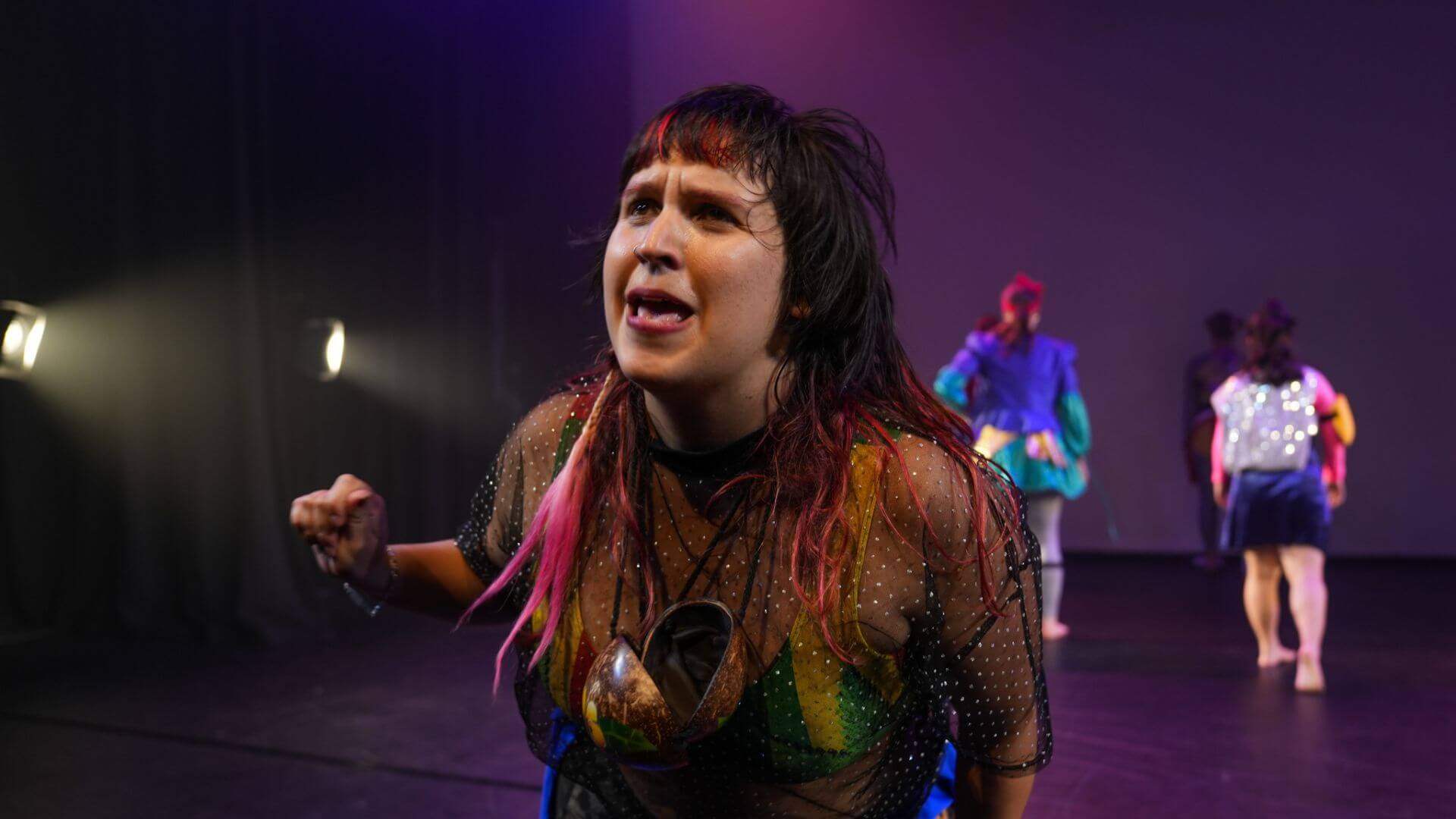 an image of a person on a mauve-lit stage, speaking expressively to the audience, wearing a coconut shell as a necklace and brightly coloured swimmers underneath a sheer top.