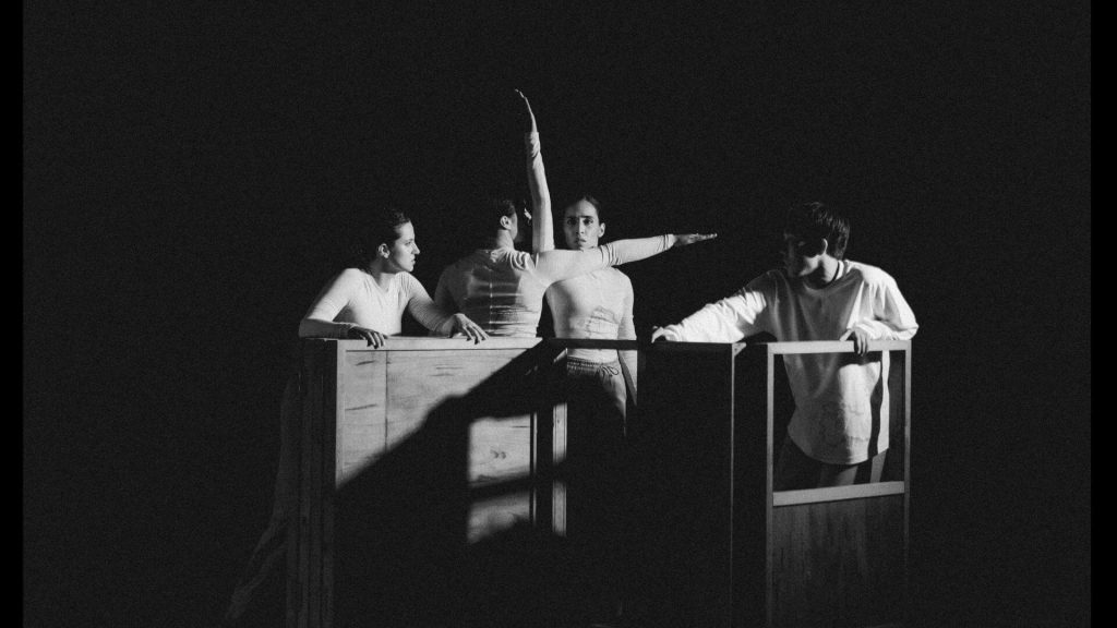 An image of four dancers performing inside a small wooden construction that appears to be a shipping crate deconstructed. Two performers have an arm extended, one vertically and one horizontally.