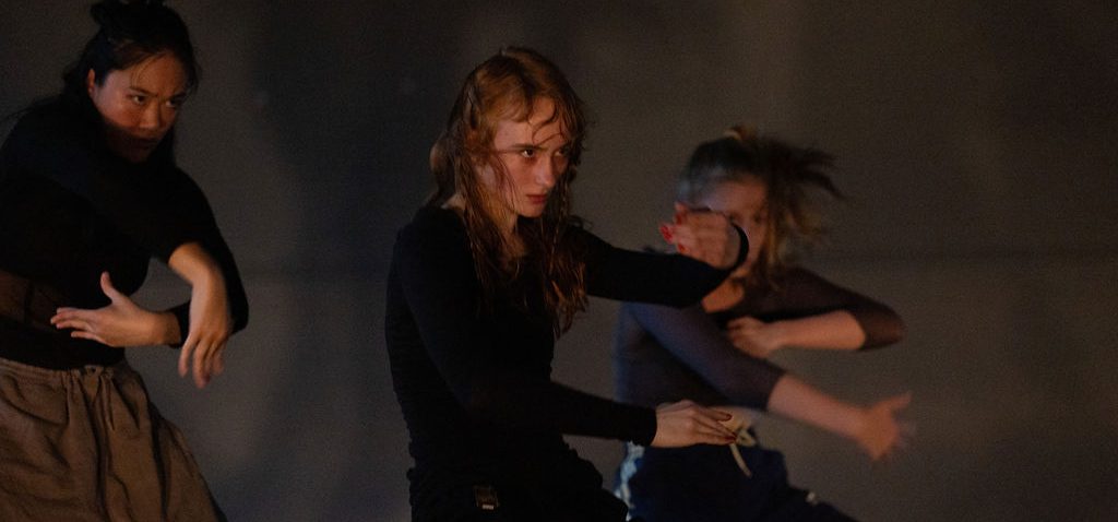 Photograph of three dancers in a rounded shape staring at the same point. They wear dark clothes and are photographed in front of a dark grey wall with their shadows enlarged behind.