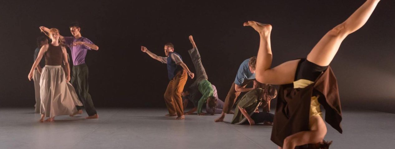 An image of a group of ten dancers performing in colourful clothes. Many of the dancers are in contact with each other and one in the forefront of the space is balancing in a headstand.
