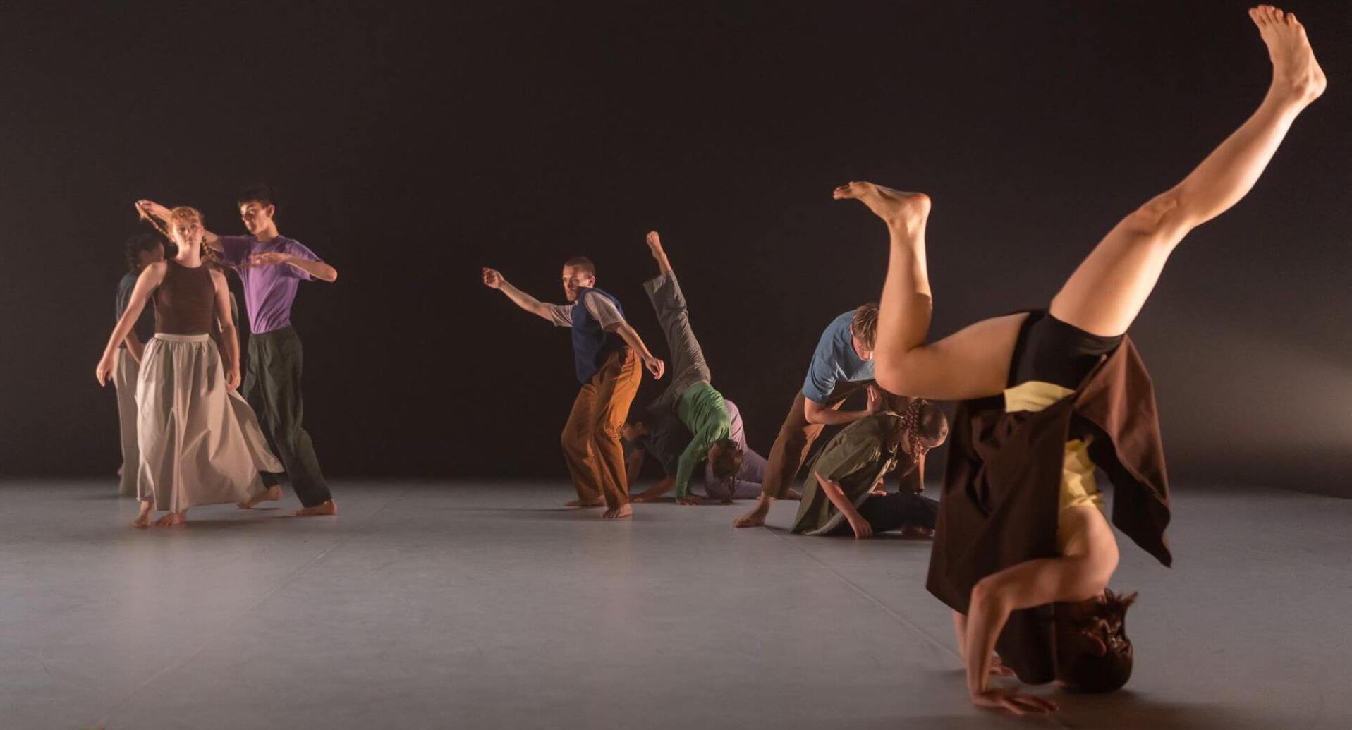 An image of a group of ten dancers performing in colourful clothes. Many of the dancers are in contact with each other and one in the forefront of the space is balancing in a headstand.