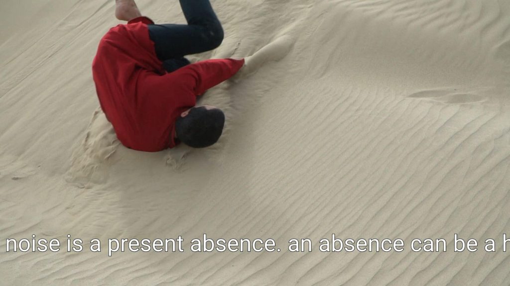Still of a male figure dressed in a bright red kurta rolling down a sand dune. He is tumbling over his right shoulder and is sliding down the dune. Subtitles at the bottom of the image read “noise is a present absence. An absence can be a h-“. The rest of the text is cut off.