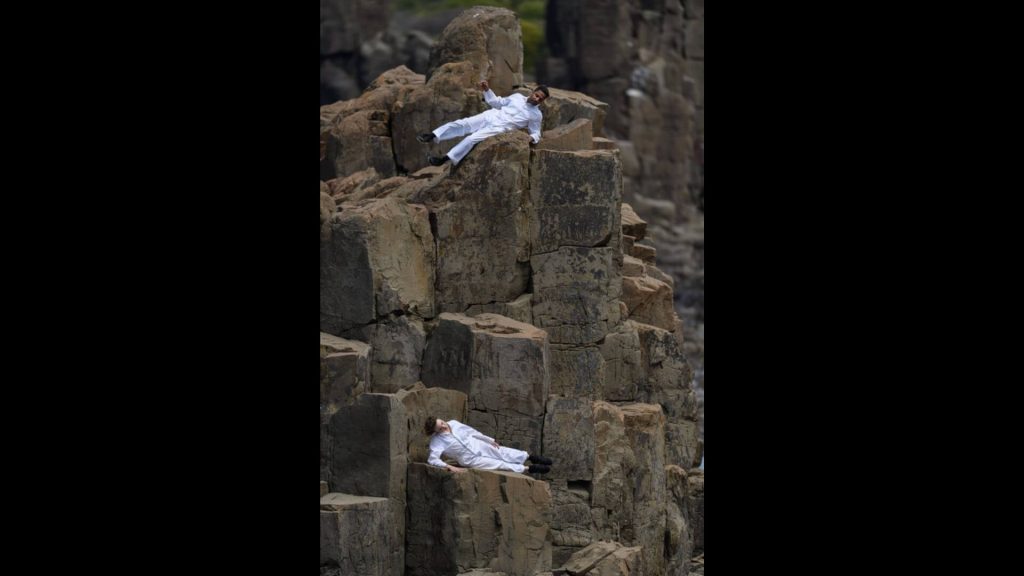 An image of a striking rock formation with two performers perched on different parts of the rock, one several metres above the other one. Both performers wear white overalls that stand out brightly against the brown rock.