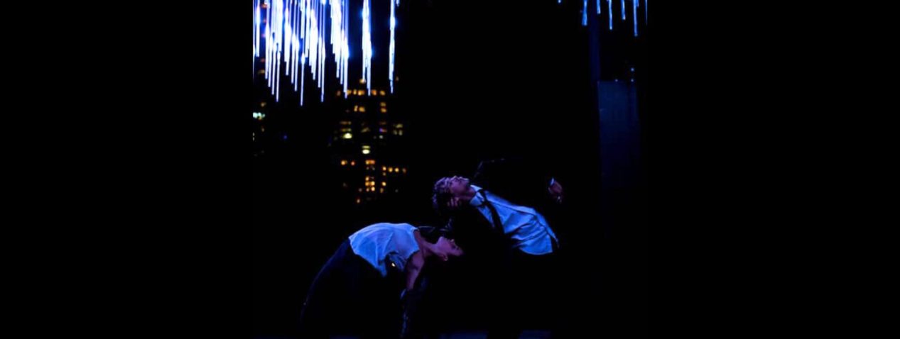 Two performers facing away from each other arch their spines to bend backwards towards each other. Dressed in suits, they stand amidst cityscape with spectacular lights.