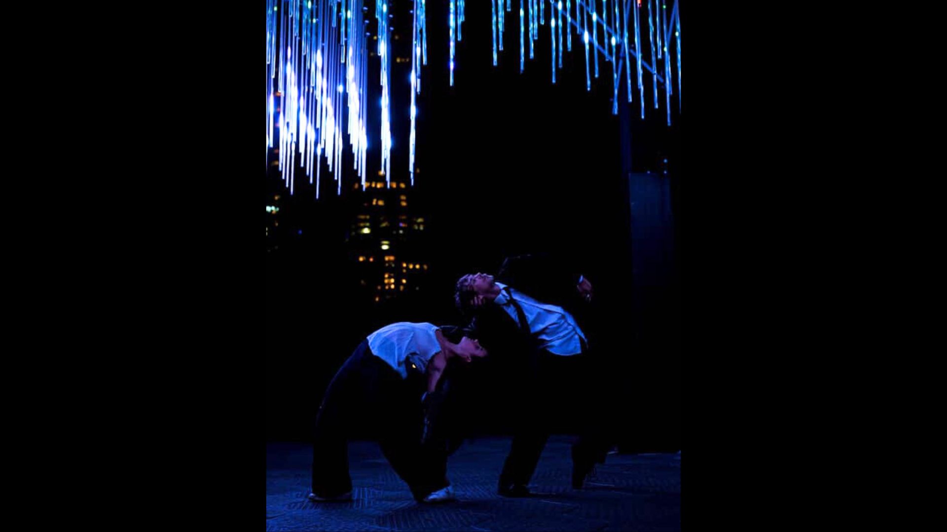 Two performers facing away from each other arch their spines to bend backwards towards each other. Dressed in suits, they stand amidst cityscape with spectacular lights.