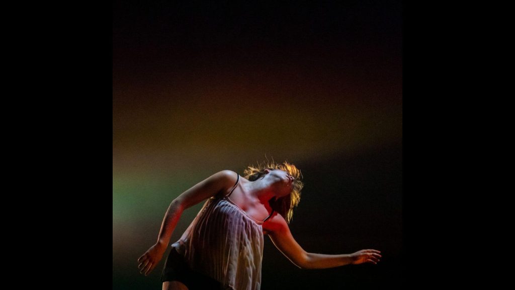 An image of a dancer in motion with a sense of abandonment, their head tilted back and hair falling out behind them. They are lit by by warm theatrical lighting.