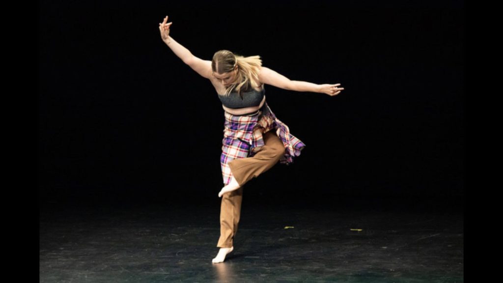 Photograph of a female figure with her right arm raised upwards and her left arm to the side. Her fingers are grouped together, suggesting stag antlers, and her left foot is raised towards her opposite leg, with her knee turned out. She wears a grey sports crop top, a purple tartan kilt, and brown suit pants.