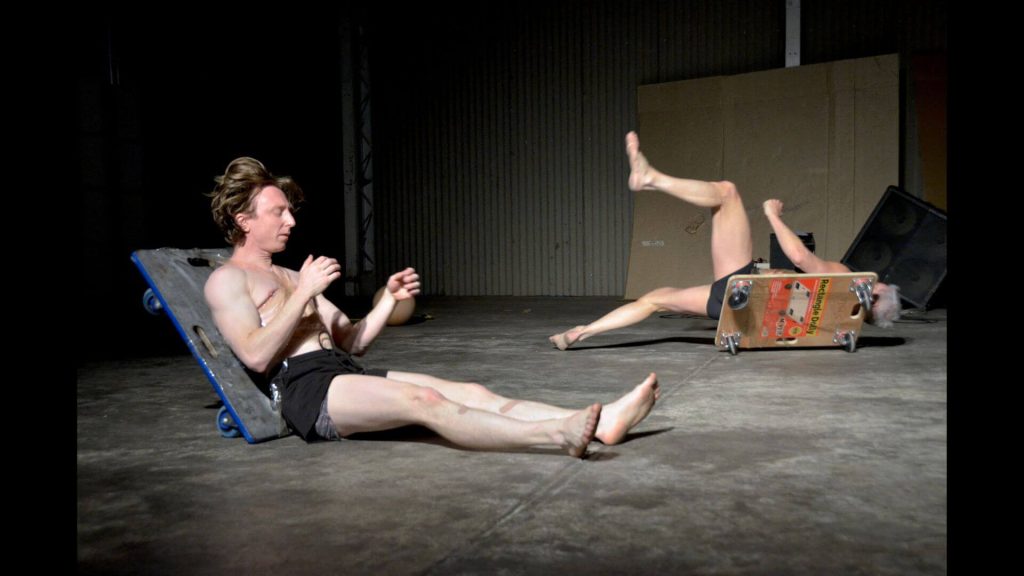 Photograph of two persons on a concrete floor in black shorts with dollies sticky taped to their backs falling over in an ungraceful way.
