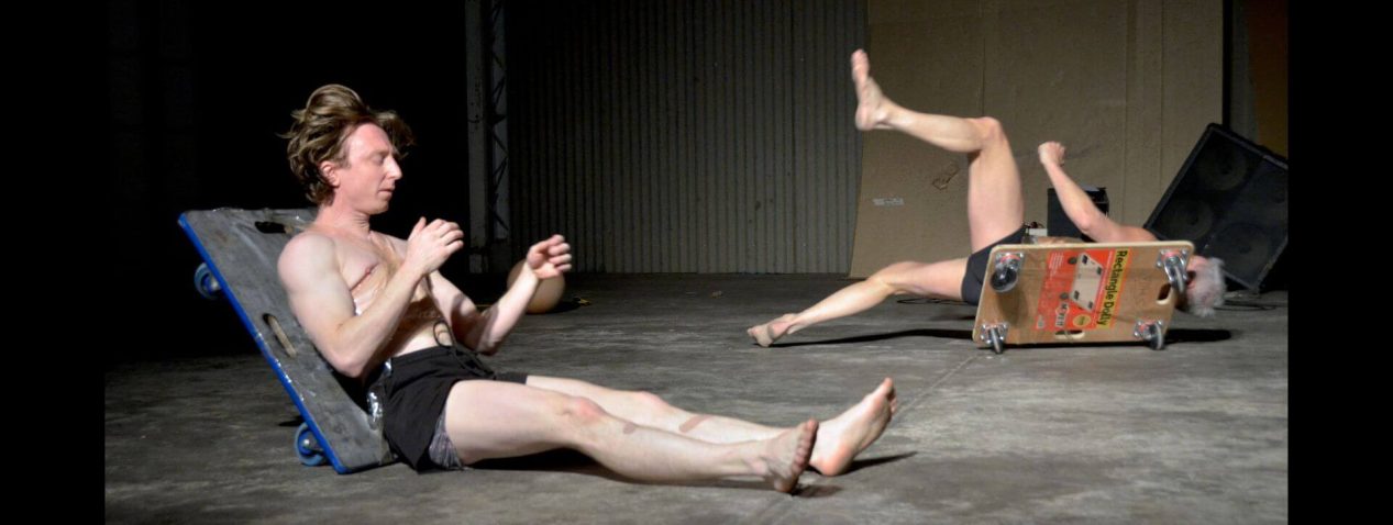 Photograph of two persons on a concrete floor in black shorts with dollies sticky taped to their backs falling over in an ungraceful way.