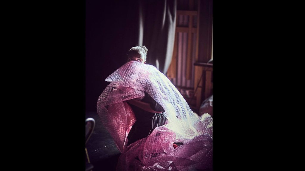Photograph of a dancer kneeling and looking over the shoulder. Red bubble wrap is draped over the shoulder and partially covers the face; the light from an adjacent window pours over the dancer’s back.