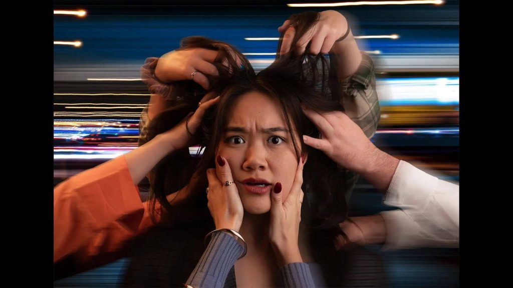 A woman looks into the camera with a confused expression. Hands around her head pull at her hair and distort her face. In the background the world rushes by.