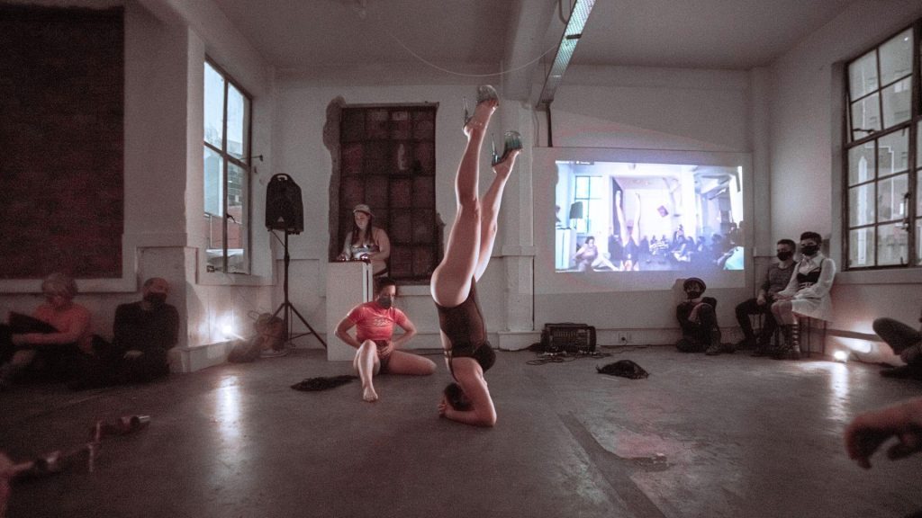 Photograph of a woman doing a headstand wearing a black bodysuit and stripper shoes. Behind her is another performer in a sitting position and a musician standing behind a plinth playing with an electronic soundboard, both are also in underwear. In the background is a video projection of the scene and audience are sitting around the edge of the room watching.