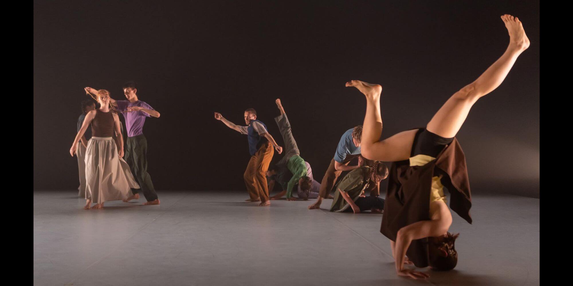 An image of a group of ten dancers performing in colourful clothes. Many of the dancers are in contact with each other and one in the forefront of the space is balancing in a headstand.