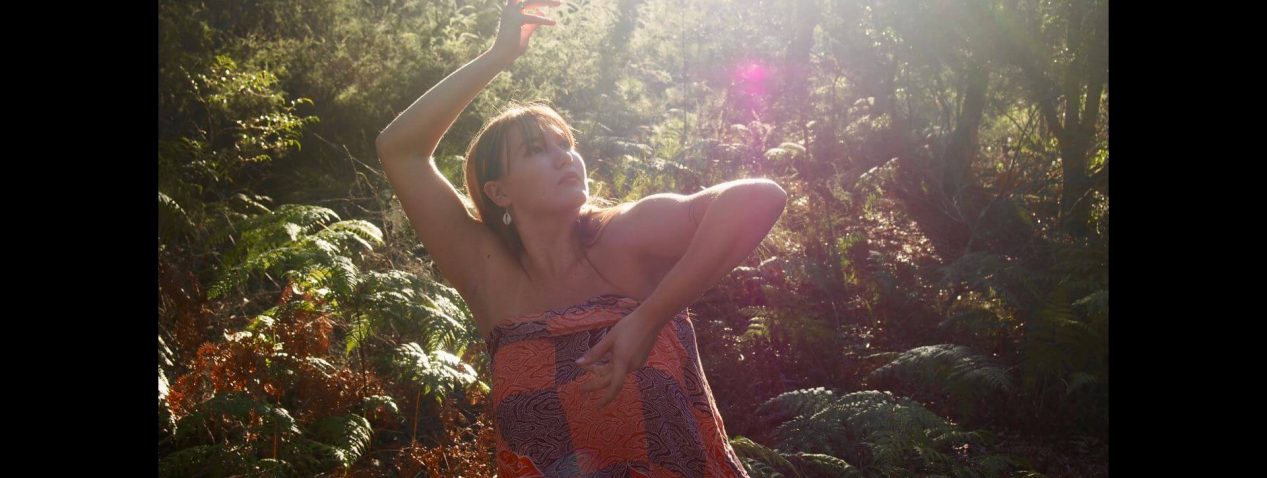 Portrait of person dancing with arms folded around their face, looking up, outside in a rainforest with many green ferns.