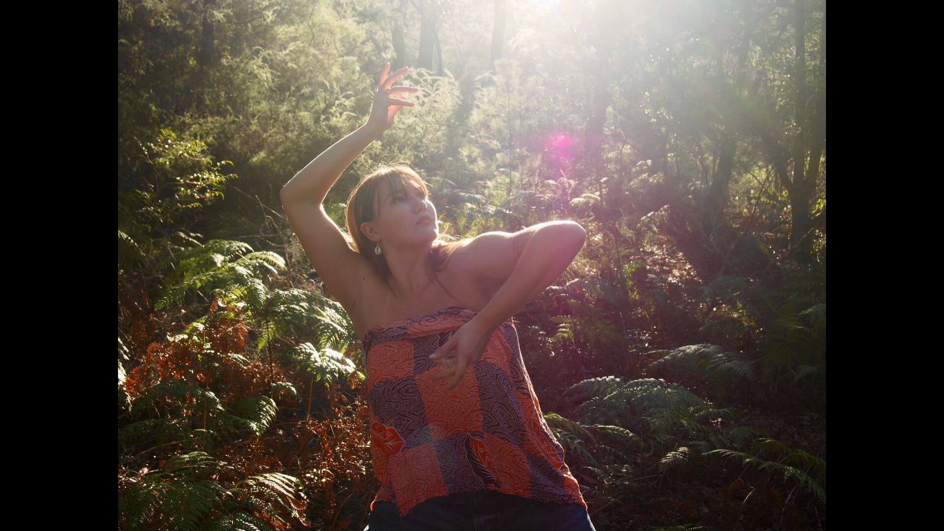 Portrait of person dancing with arms folded around their face, looking up, outside in a rainforest with many green ferns.