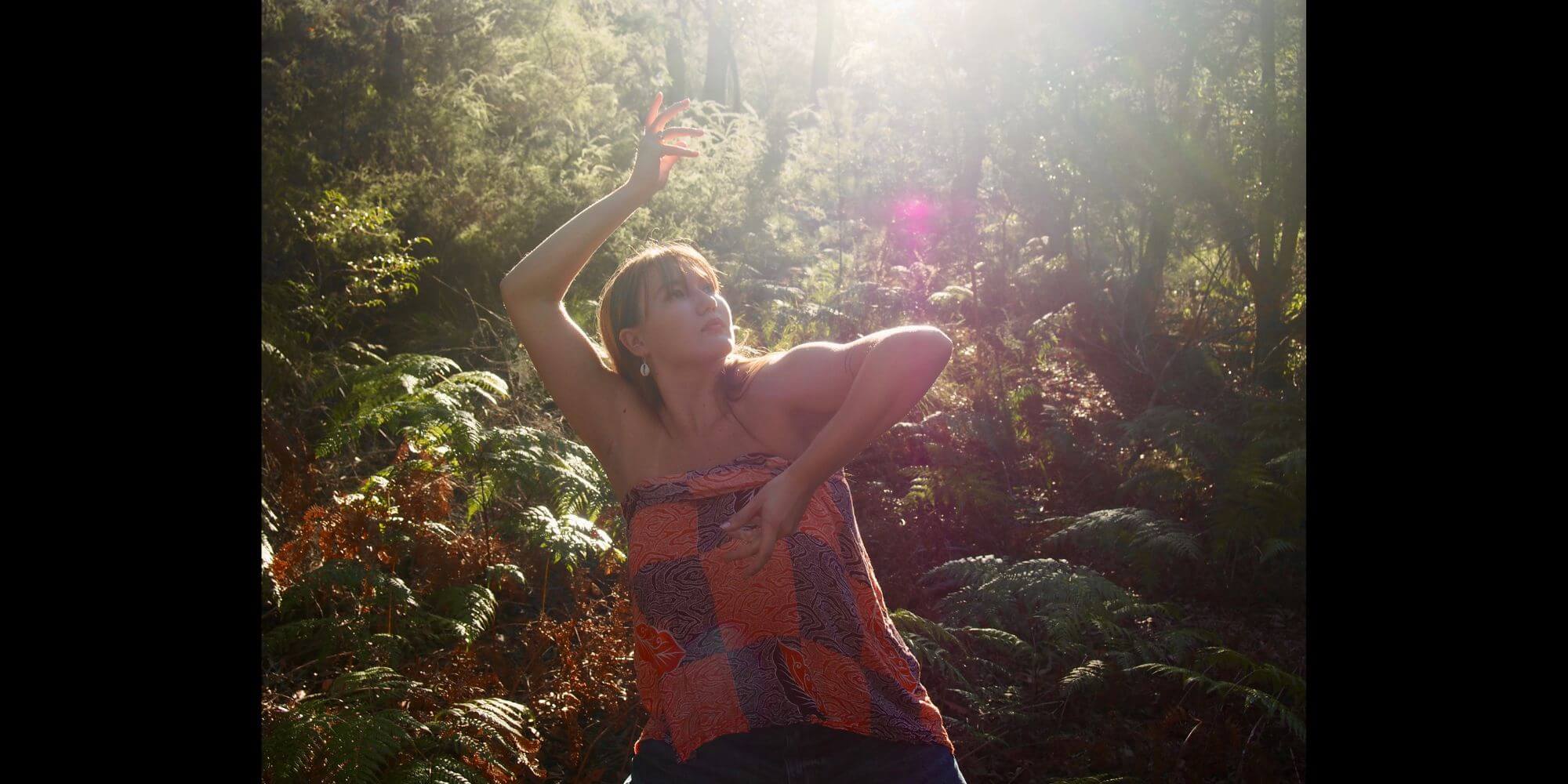 Portrait of person dancing with arms folded around their face, looking up, outside in a rainforest with many green ferns.