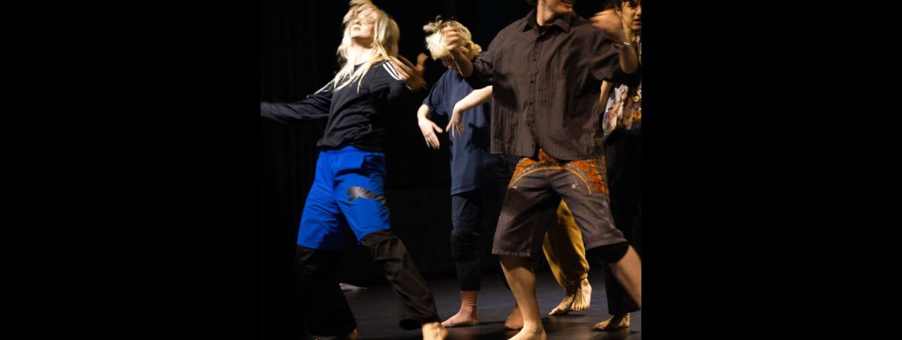 A photo of a group of performers standing close together performing in a showing. One is looking up with arms opened wide, one is looking down with arms and wrists folded in towards their belly, one is looking behind them with a large smile across their face.