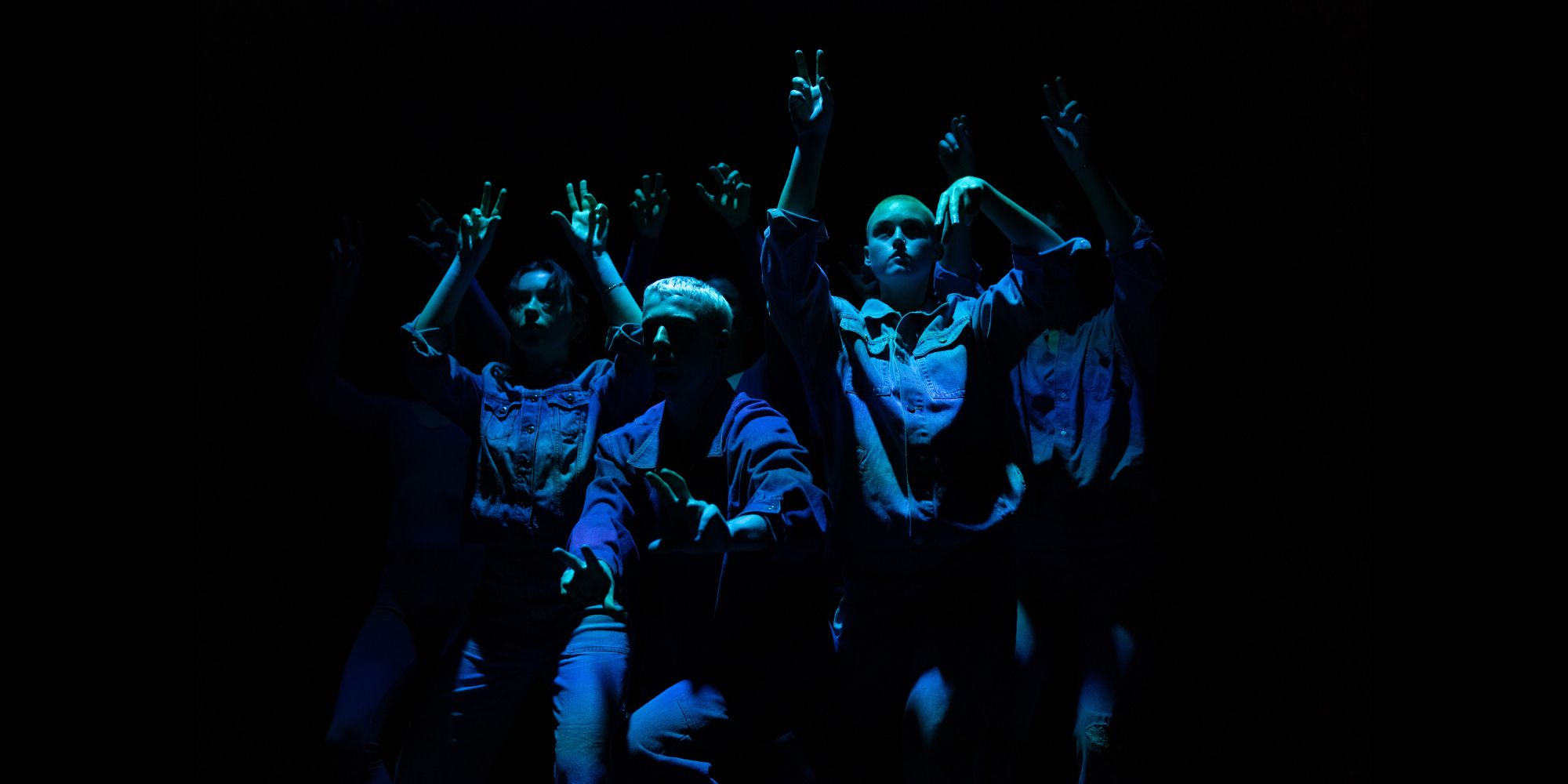 Image of a group of dancers performing with fingers curling like animal paws, wearing blue denim and lit with an indigo hue.
