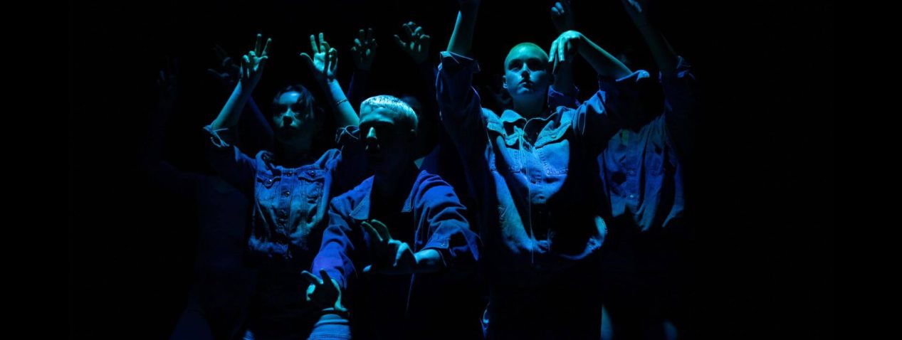 Image of a group of dancers performing with fingers curling like animal paws, wearing blue denim and lit with an indigo hue.