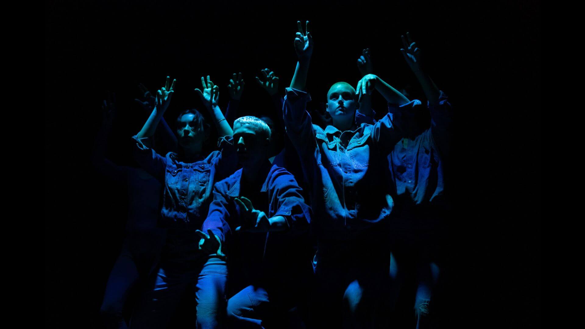 Image of a group of dancers performing with fingers curling like animal paws, wearing blue denim and lit with an indigo hue.