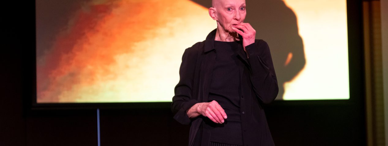 Image of a person performing with a bright projection of orange hues behind them on the backdrop. The performer has their fingers close to their mouth and is looking with wide eyes directly into the camera.