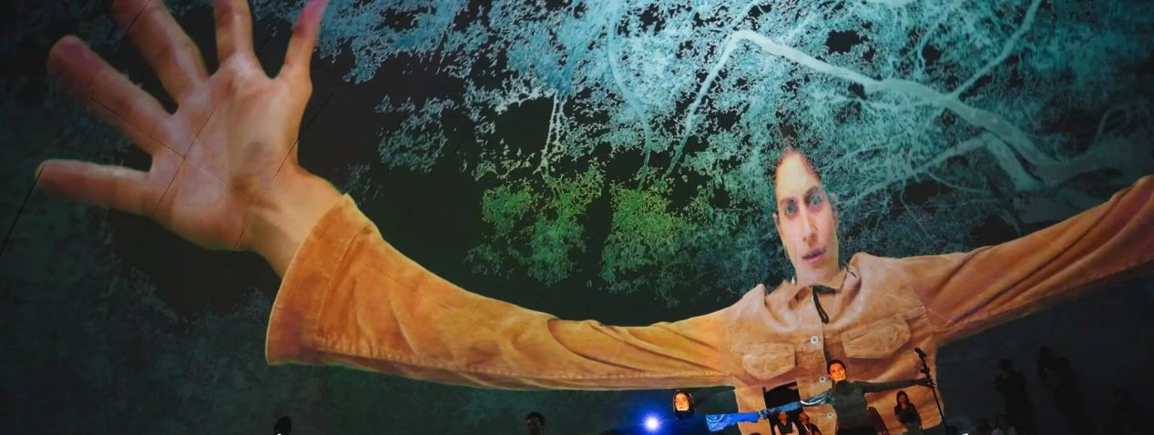 Three performers stand side by side with arms stretched out towards each other. Behind them is a large projection of one of the performers with arms and hands stretched out and magnified to a more-than-human scale.
