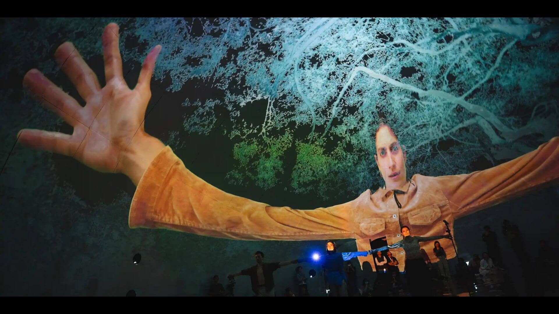 Three performers stand side by side with arms stretched out towards each other. Behind them is a large projection of one of the performers with arms and hands stretched out and magnified to a more-than-human scale.